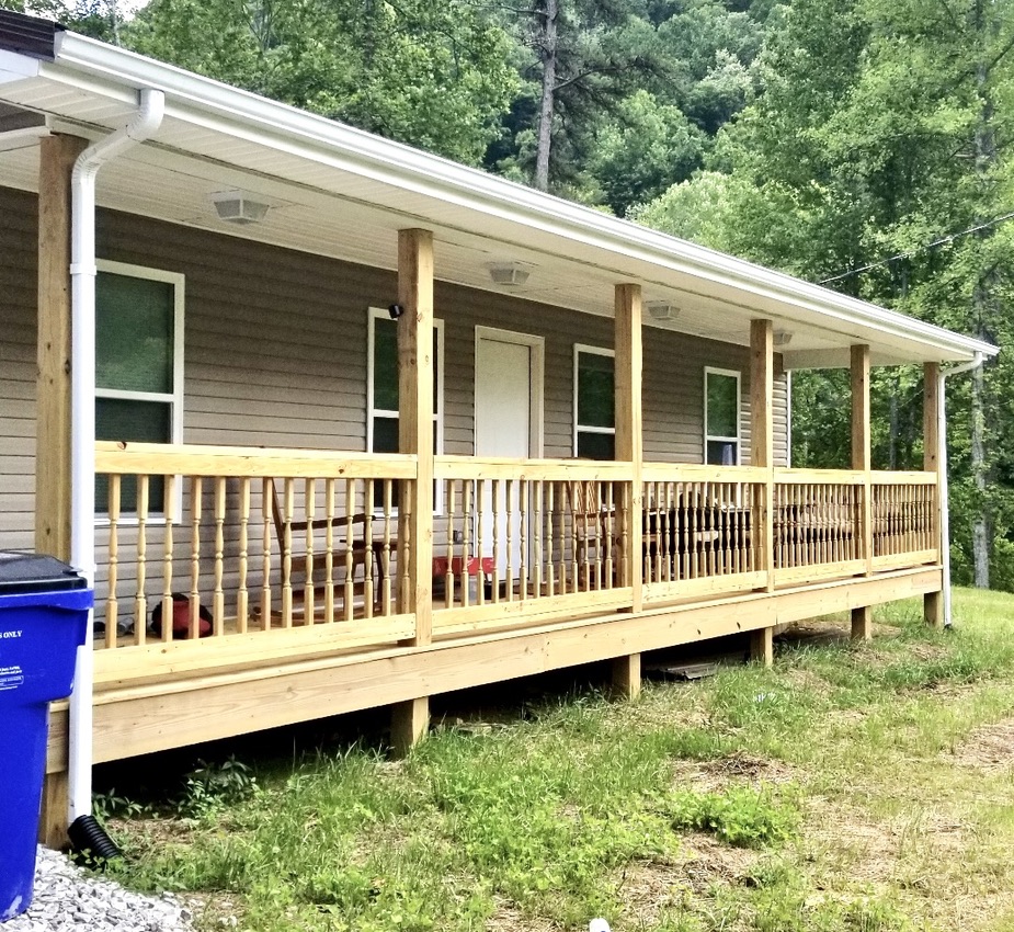 Photo shows the front of a newly built house with tan siding and a wood porch. A lawn is seen in front of it and trees behind it.