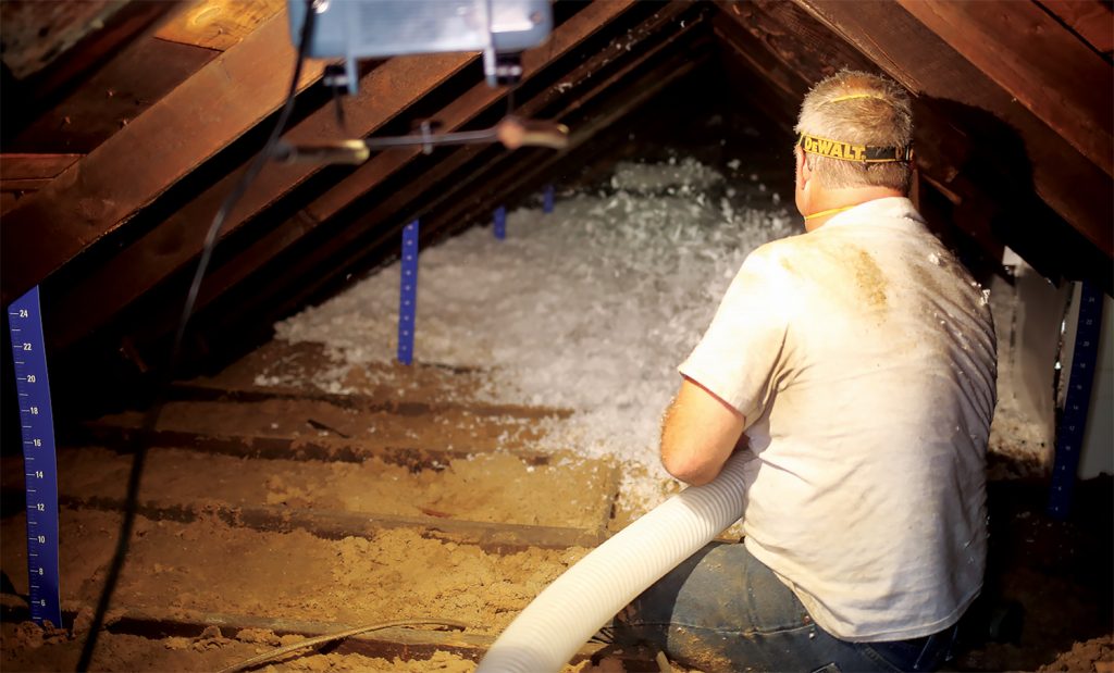 A man in a white shirt with his back to the camera uses a flexible tube to blow insulation into an attic.