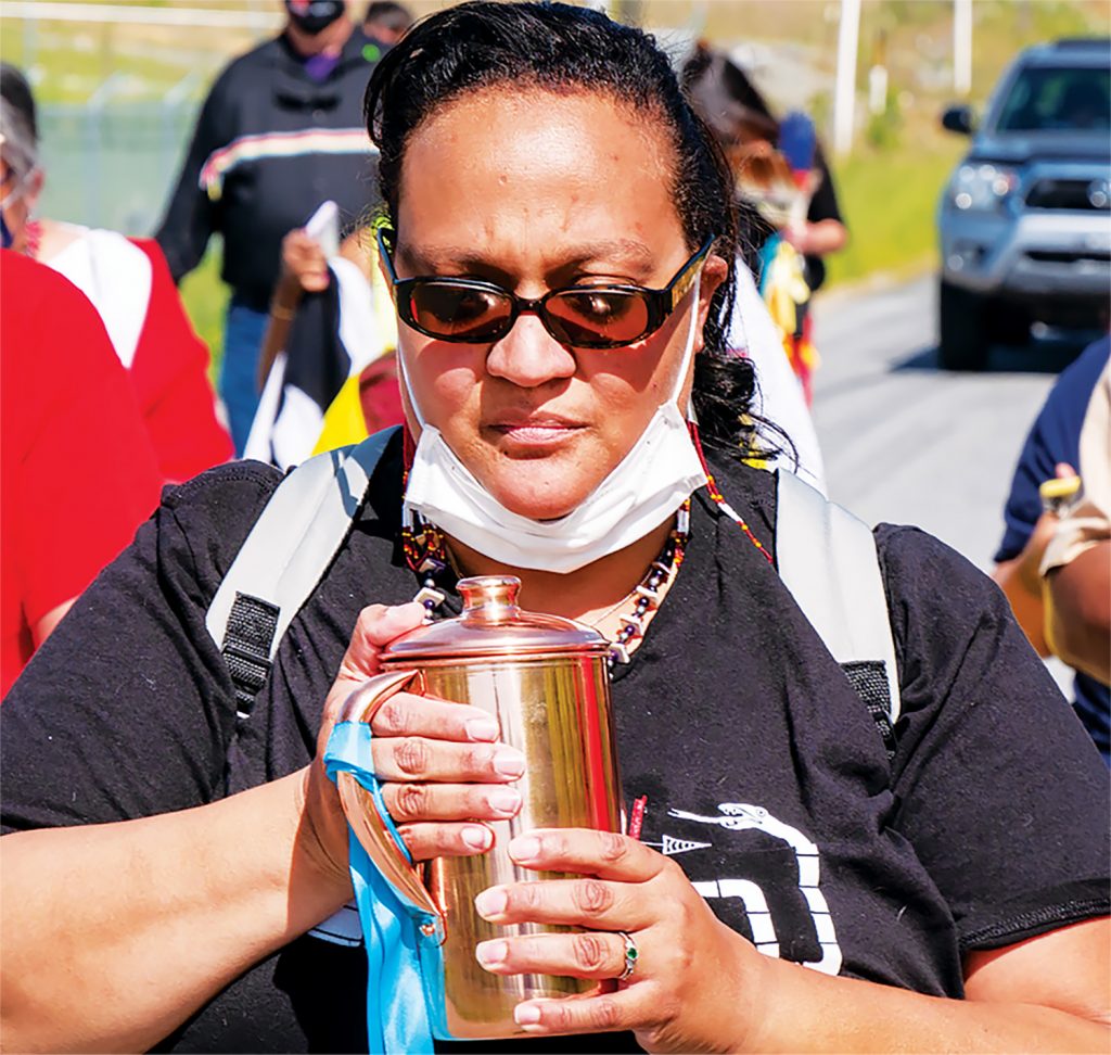 An Indigenous woman holds a vessel of water as she walks down a road in a group of people.