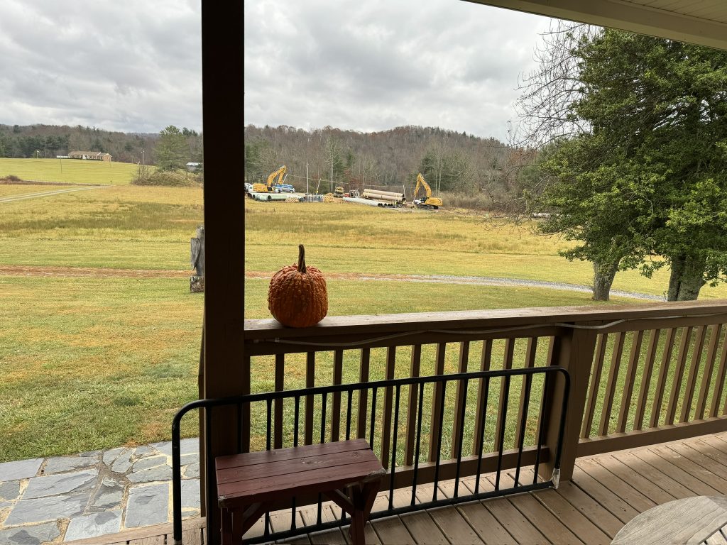 A pumpkin sits on a porch rail. Across the lawn from the porch is a large construction staging area with cranes and lengths of huge pipe visible.