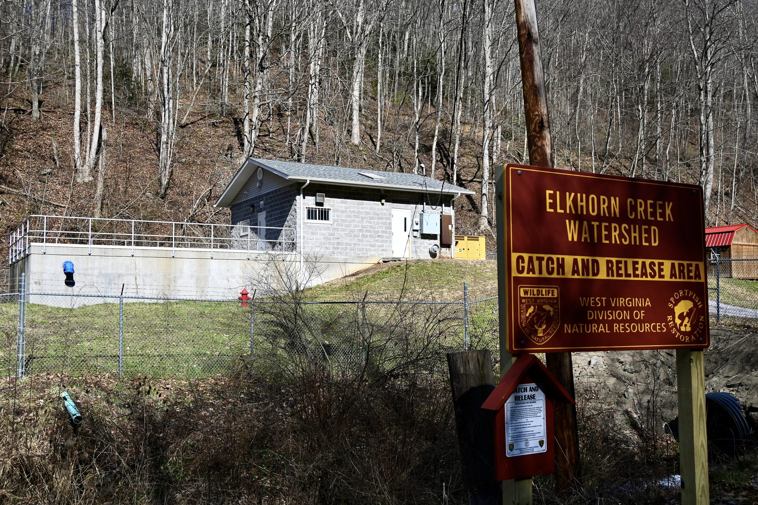 A cement block building sits behind a fence with barbed wire across the top. A sign in the foreground says "Elkhorn Creek Watershed, Catch and Release Area, West Virginia Division of Natural Resources."