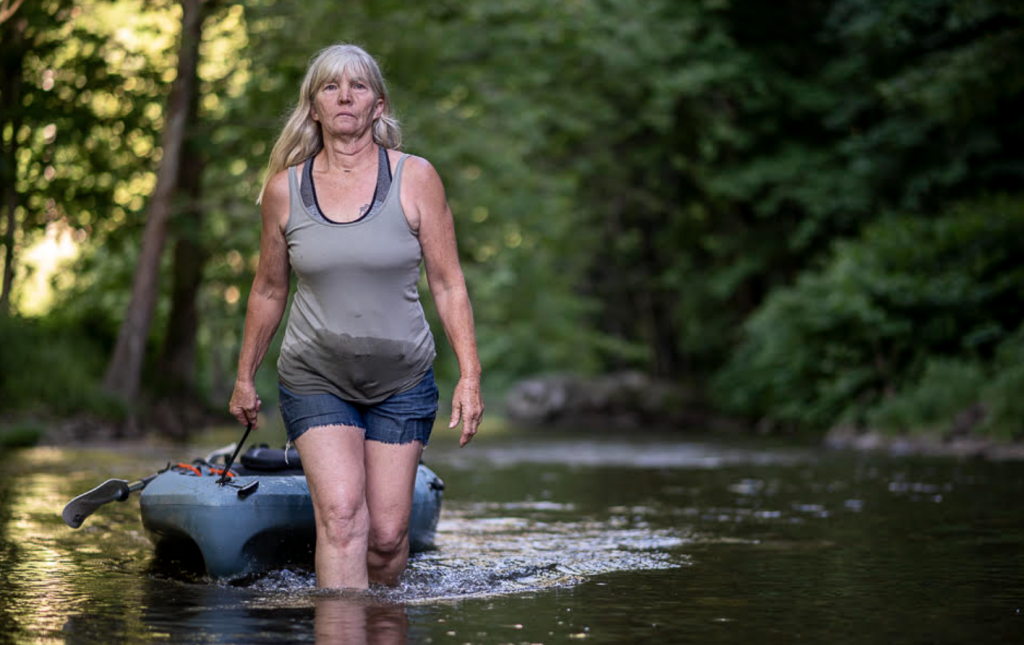 A woman with long gray hair pulls her kayak as she walks through a shallow river