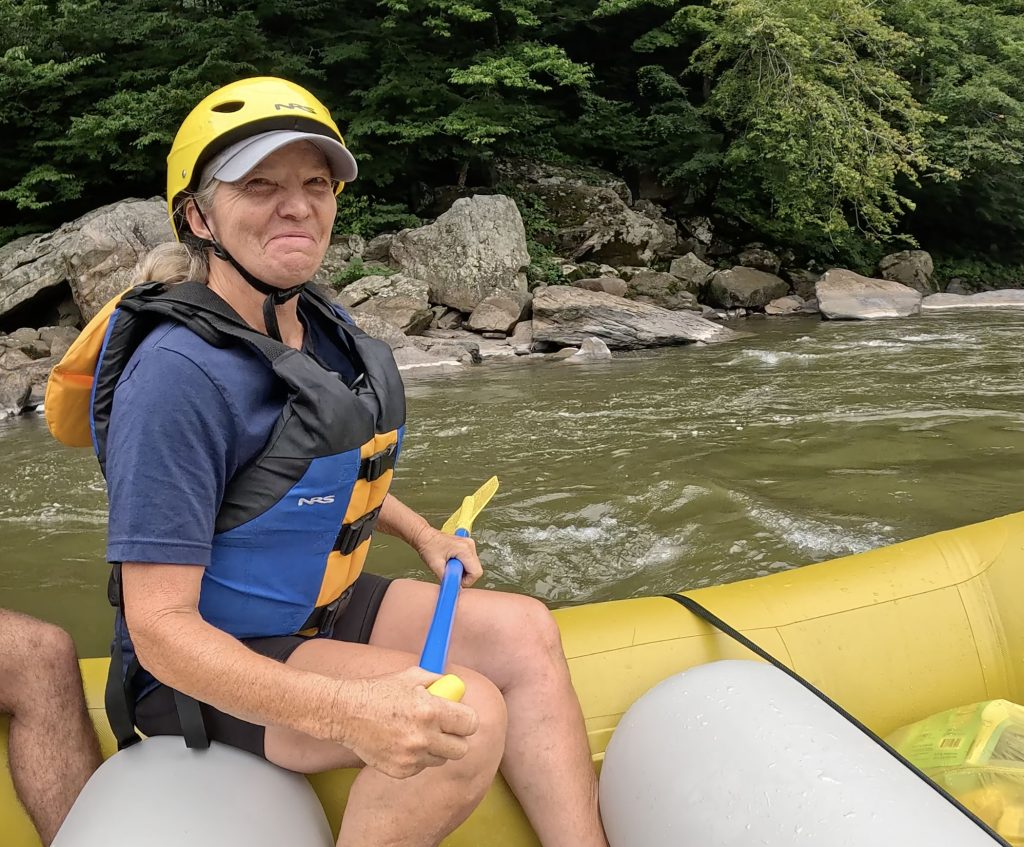 A woman smiles as she sits on a raft in a river holding a paddle.