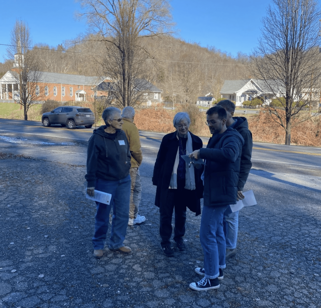 A group of people stand gathered on a street with the town of Pound visible in the background.