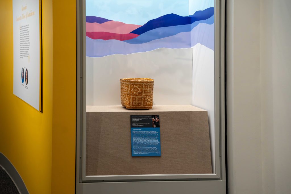 A woven basket sites behind a display case in a museum. “I am a generational weaver, learning from my mother. My grandmother and great grandfather were beasketweavers too,” writes artist Vivian Garner Cottrell in her artist statement about her traditional, Southeastern Woodlands style doublewoven basket, “The Sun Dances Around the People.” Photo by Abby Hassler