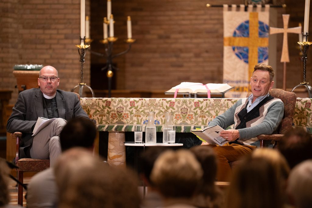 A man, Silas House, sits beside a male priest during a Q&A about All These Ghosts put on by the Episcopal Diocese of East Tennessee