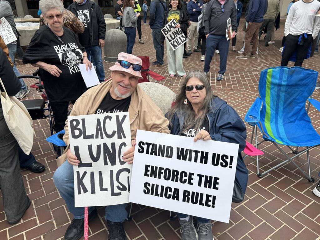 two people sit outdoors with signs: one sign says "black lung kills" and the other says "stand with us: enforce the silica rule"
