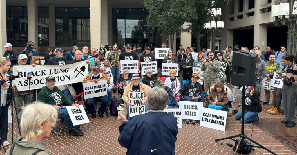 A large group of people gathered outside a civic building at a protest, many holding signs reading “Black Lung Kills,” and “Silica Kills."