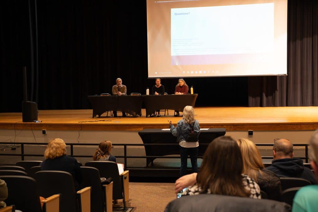 Woman addresses panel of three experts on stage in high school auditorium