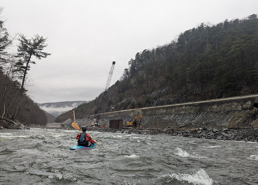 A man kayaks on a rainy, cloudy day in choppy waters by ongoing interstate construction