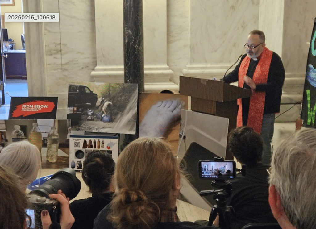 Pastor Brad Davis speaks at a podium inside the West Virginia State Capitol, addressing a small audience. In front of him, photos and displays show images of pollution, contaminated water, and environmental impacts, while attendees watch and record the presentation with cameras and a smartphone.