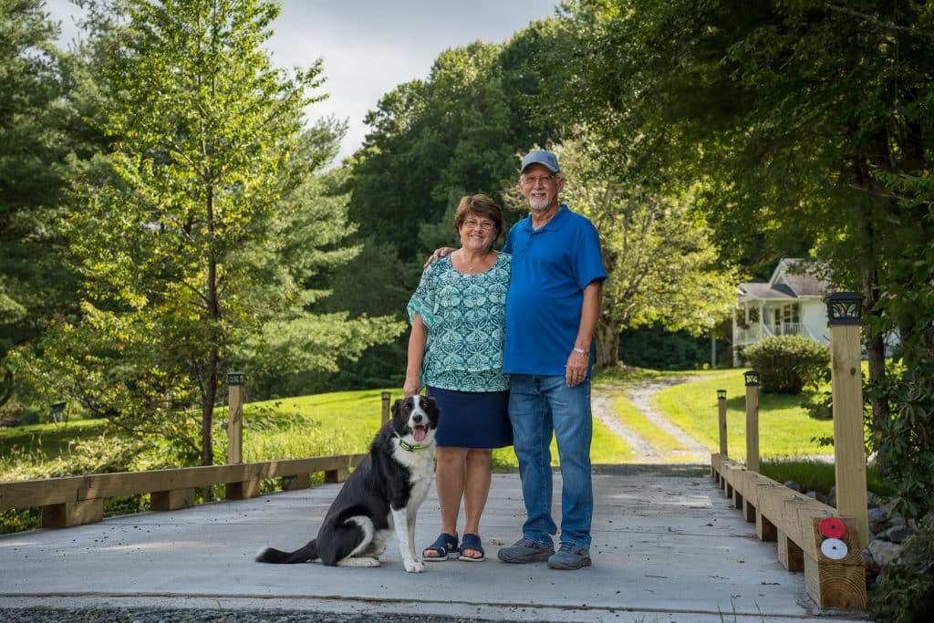 Alice and Gary Stevens stand on their new 40-foot bridge with their dog in Watauga County, N.C.