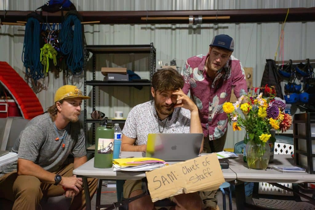 Three men crowd around a laptop on a foldout table in a warehouse that also holds a cardboard sign that reads "Sam: Chief of Staff" and a vase of flowers