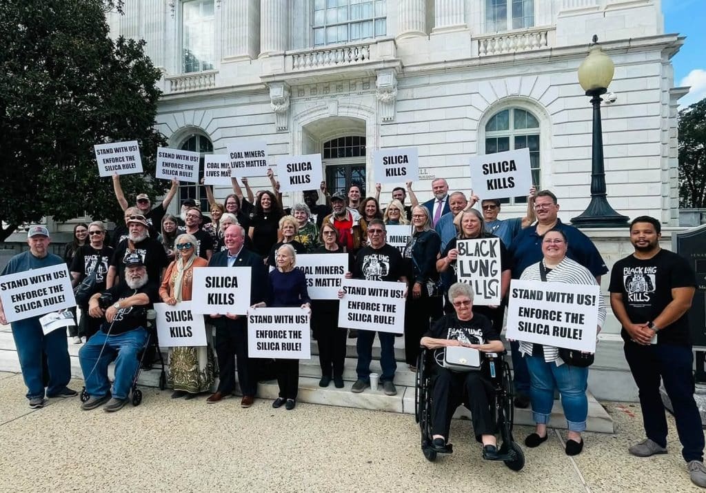 Several dozen miners and their supporters wearing "Black Lung Kills" t-shirts and carrying signs stand on the steps of a capitol building