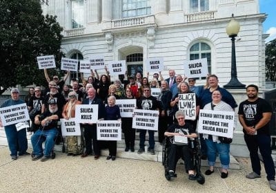 Several dozen miners and their supporters wearing "Black Lung Kills" t-shirts and carrying signs stand on the steps of a capitol building