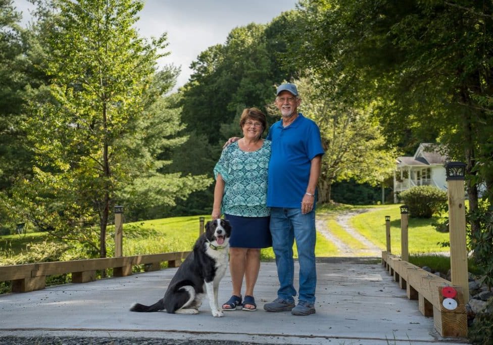 Alice and Gary Stevens stand on their new 40-foot bridge with their dog in Watauga County, N.C.