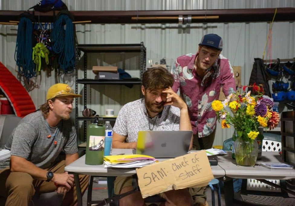 Three men crowd around a laptop on a foldout table in a warehouse that also holds a cardboard sign that reads "Sam: Chief of Staff" and a vase of flowers